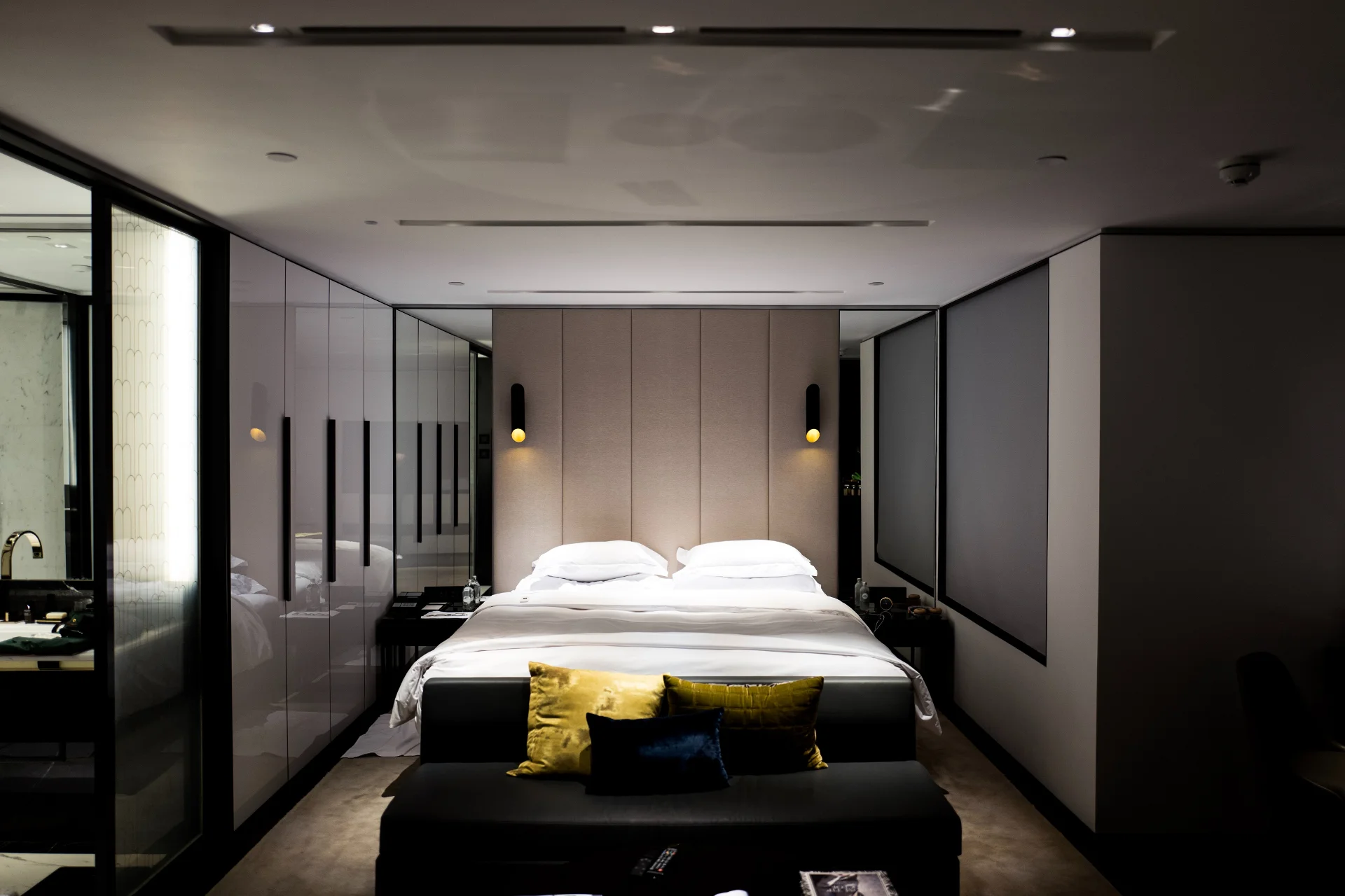 Dans un design épuré, un plafond blanc laqué avec des spots encastrés, des murs gris clair et une tête de lit taupe avec deux lampes de chevet fixées à chaque bout de la tête de lit. Un lit au centre avec une literie blanche et un banc coffre en bout de lit noir avec des coussins jaunes. A gauche de la photo, on voit derrière un panneau coulissant une salle de bain. Cette photo n’est pas contractuelle et n’est là que pour illustrer le standing de chambre visé.