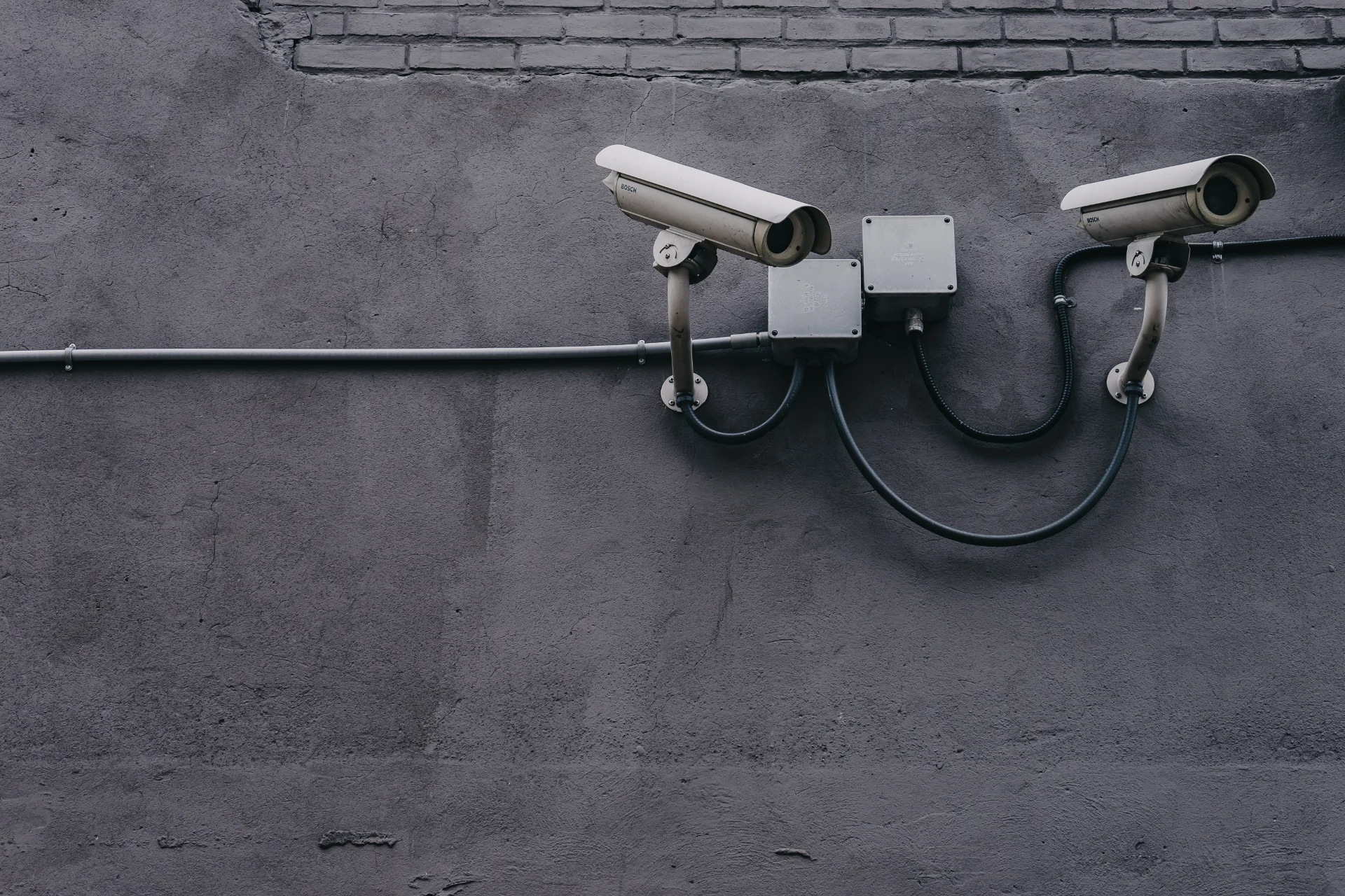 Photo dans les tons gris cendrés avec deux caméras de surveillance blanche fixées au mur dont on voit quelques briques.